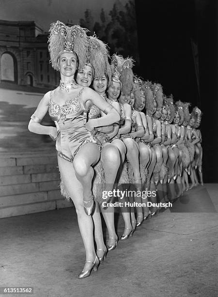 The showgirls, the Tiller Girls pose in a line wearing costumes with feather headdresses.