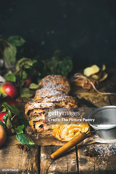 apple strudel cake cut in pieces with fresh red apples, sugar powder and spices on rustic wooden table - strudel dessert stock-fotos und bilder