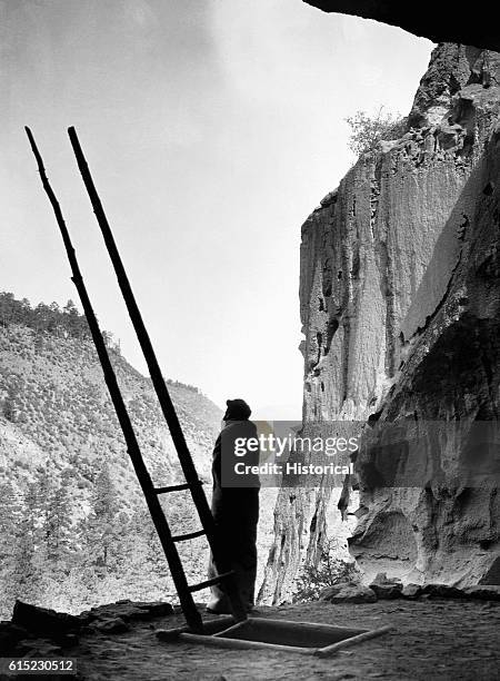 Man stands next to the entrance of a kiva and its wooden ladder, among the restored ruins of a Native American cliff dwelling at Bandelier National...