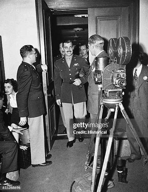 Actor Clark Gable walks into a room, wearing a military uniform. A movie camera is set up to film his appearance.