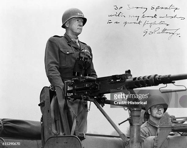 George Patton , American general, stands on a military vehicle in front of an artillery gun.
