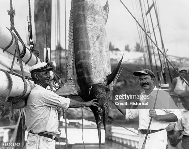 Ernest Hemingway stands with a fishing rod and a marlin, while Captain Joe Russell from Key West looks on. Hemingway , American writer, is known for...