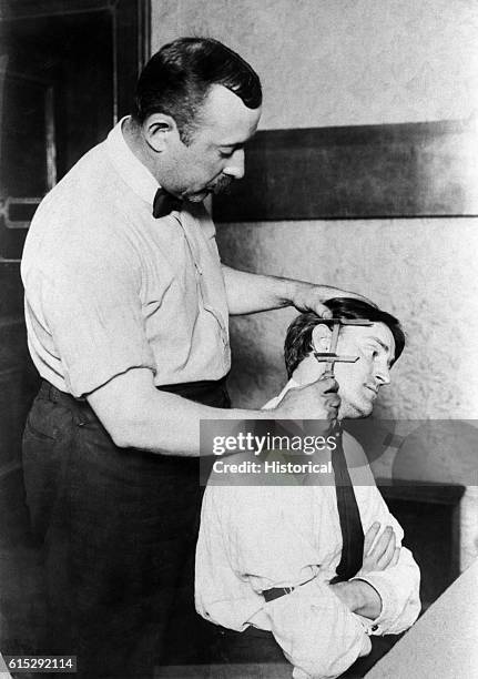 Police officer takes Bertillon measurements of a suspect's[?] ear at New York Police Headquarters. The Bertillon system is a series of physical...