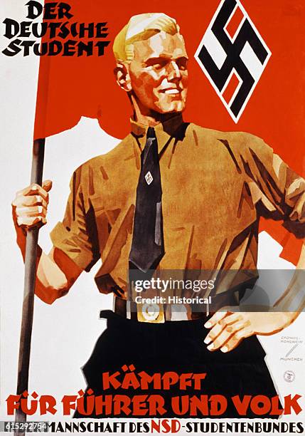 Young blond man wearing a military uniform supports a nazi flag with one hand, the other hand on his hip. He stands upright and smiles.