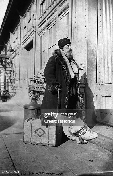 An elderly Russian Jewish immigrant stands outside a building on Ellis Island, wearing heavy clothing and surrounded by all his luggage. New York,...