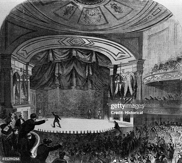 Actor and assassin John Wilkes Booth runs across the stage of Ford's Theater after having shot President Abraham Lincoln. A man climbs up on stage to...