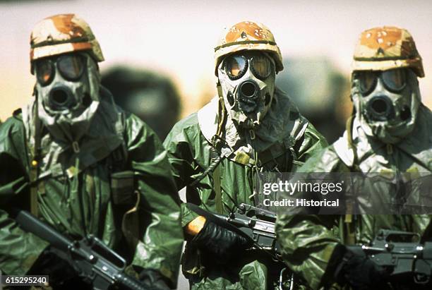 Dressed in rain suits, gloves and M-17A1 protective masks, three soldiers from the 82nd Airborne Division walk around their camp to acclimate their...