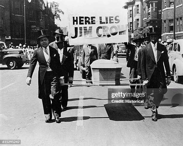 African-American men wearing tuxedos carry a coffin and a "Here Lies Jim Crow" sign down the middle of a street as a demonstration against "Jim Crow"...