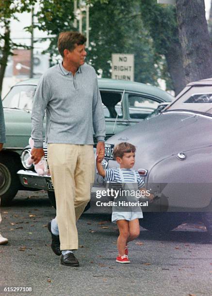 John F. Kennedy walks holding John Jr.'s hand at Hyannisport, Massachusetts. The president carries a pink teddy bear, and John Jr. Carries a toy...