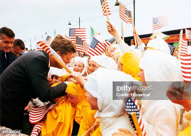 President Kennedy is greeted by Irish school children in Galway, Ireland. The children, dressed in white or gold uniforms, wave American flags.