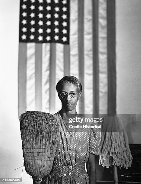 Mrs Ella Watson, a charwoman employed in a federal office building in Washington, DC, stands in front of an American flag with her mop and broom in a...