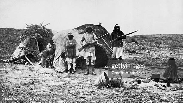 Apache rancheria with two men holding rifles. | Location: outdoors.