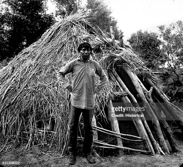 Supai Charlie, a Havasupai man, standing in front of his ha-wa, a structure made of sticks and grass. Havasu Canyon.