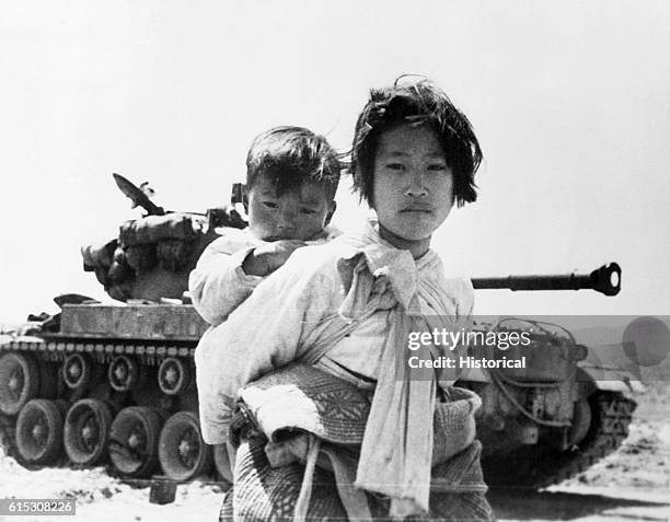 With her brother on her back, a war weary Korean girl tiredly trudges by a stalled M-26 tank, at Haenju, Korea. June 9, 1951. | Location: Haenju,...