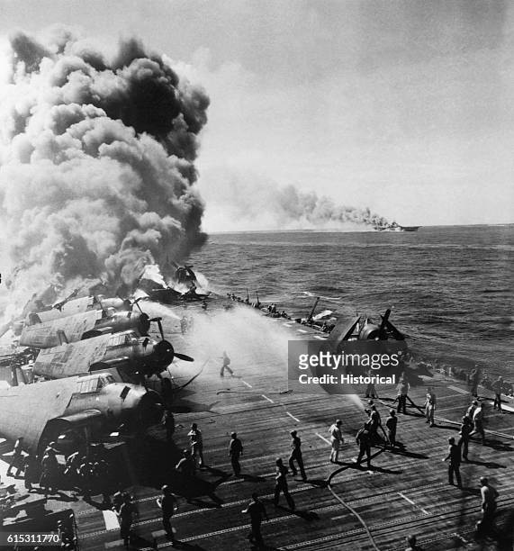 Crew members work to put out a blaze started aboard the USS Belleau Wood following a kamikaze attack. In the background, smoke pours from another...