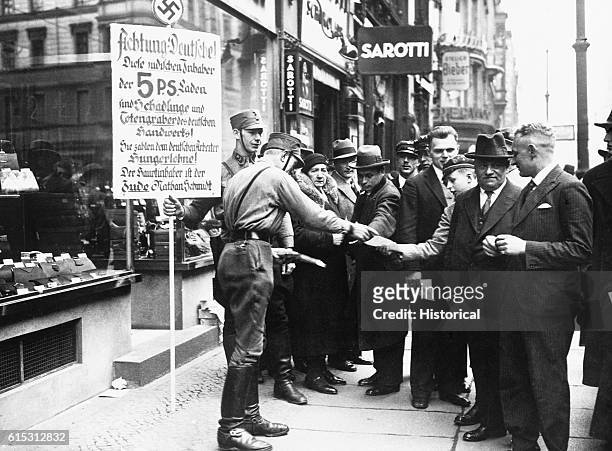 Two SA men in an unidentified German city enforce the boycott against Jewish merchants by carrying an Anti-Semitic placard in front of a store and...