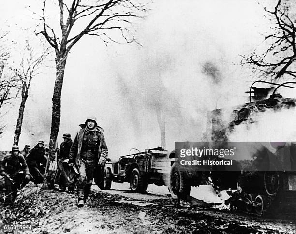 German infantry walk past burning Allied vehicles somewhere in Belgium during the Battle of the Bulge. December 1944.