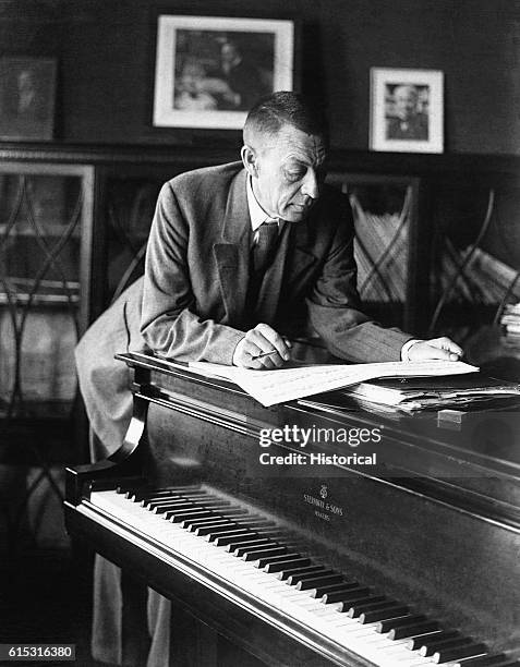 The Russian-born composer and pianist Sergei Rachmaninov reads a musical score while leaning over a piano. He is holding a cigarette in a short...