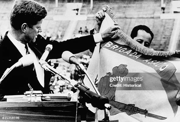 President Kennedy displays the combat flag of the Cuban landing brigade, and declares to an audience of 40,000 that it "will be returned this brigade...