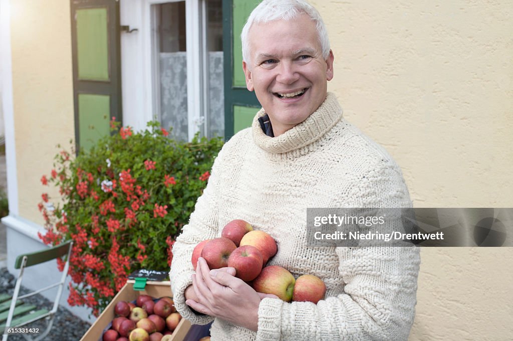 Mature man holding apples in his hands in front of wholefood shop and smiling, Bavaria, Germany
