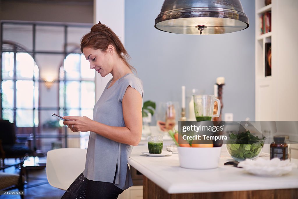 Femme détendue avec smartphone et un smoothie dans la cuisine.