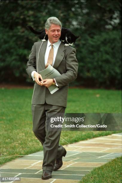 Smiling President William Jefferson Clinton wearing taupe suit walks down a stone pathway on the White House grounds with Socks the Cat, with black...