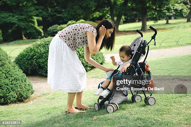 mother putting son in stroller outdoors, park on the background - baby pram in the park stock pictures, royalty-free photos & images