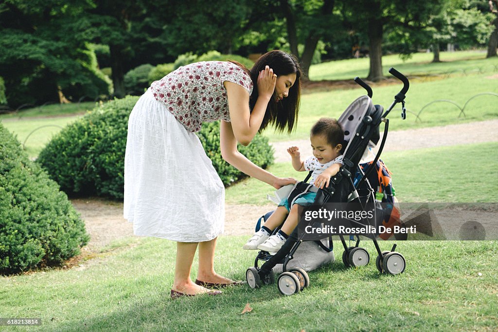 Mãe colocando filho em carrinho ao ar livre, estacionar ao fundo