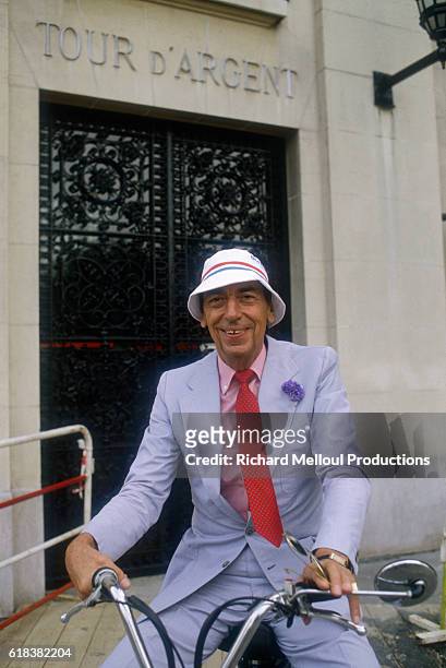 Claude Terrail sits on a moped outside his famous establishment. He is wearing a light blue suit and a beach cotton hat.