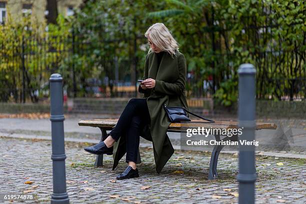 Leonie Markhorst sitting on a bench writing a text message wears a khaki green coat from Ivy + Oak, black top from Uniqlo, black skinny denim jeans...