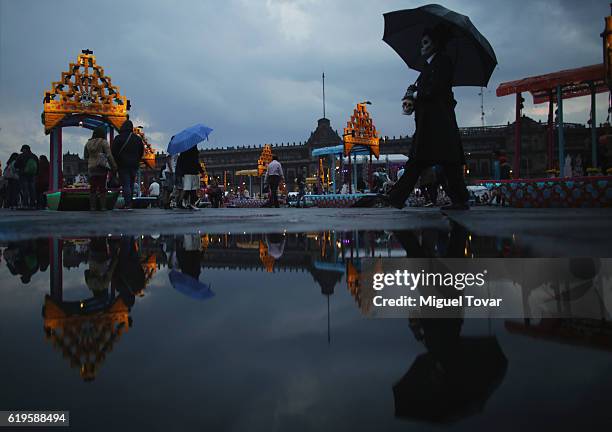Man in a costume and wearing skull makeup walks next to representations of boats known as trajineras displayed as part of the Day of the Dead...