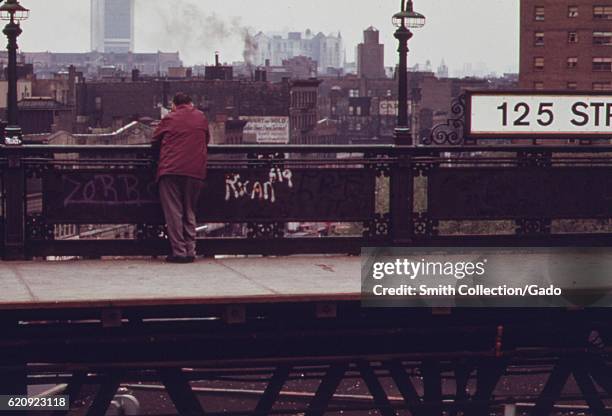 Man stands at the 125th Street Elevated Train subway platform next to several graffiti tags, New York City, New York, May, 1973. Image courtesy...
