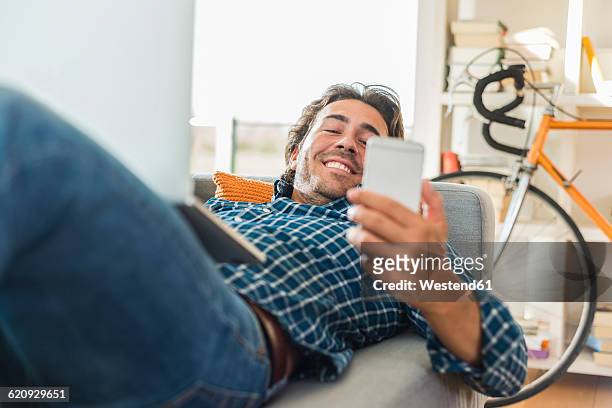 young man lying on the couch with laptop looking at his smartphone - mann handy couch stock-fotos und bilder