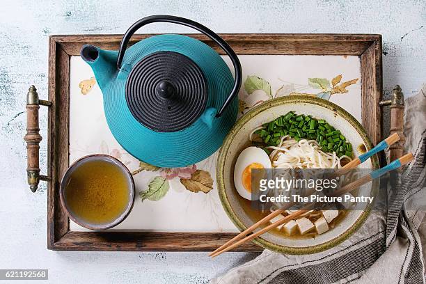 bowl of asian style soup with noodles, egg, spring onion and tofu cheese, served with chopsticks, teapot and cup of green tea on wood tray with textile over white wooden background. top view - cup noodles stock-fotos und bilder