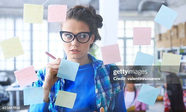 young woman writing on post-it notes on glass wall - forgot something stockfoto's en -beelden