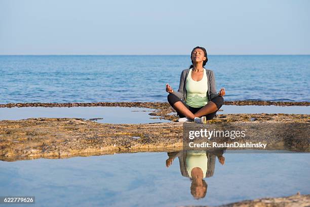 african woman meditating on seashore. - half lotus position stock pictures, royalty-free photos & images