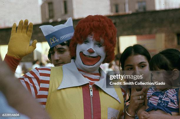 Performer dressed as Ronald McDonald waves at a camera while a Latina woman and child stand nearby and look on in the Bronx, New York City, New York,...