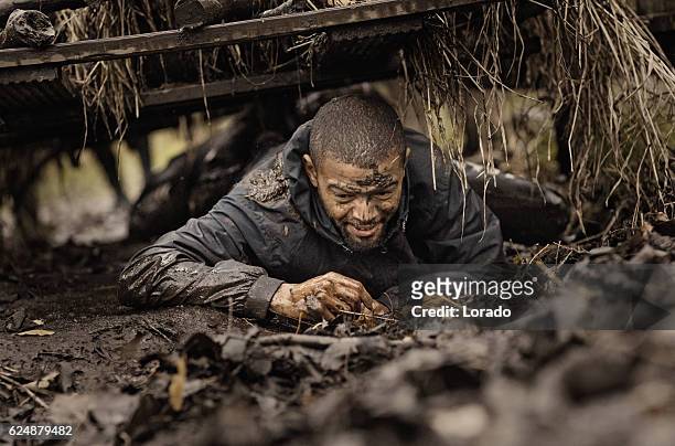 schwarz stattliche junge mann klettern unter hindernis während schlammlauf - mud run stock-fotos und bilder