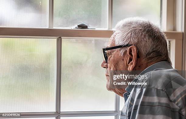 uomo anziano malincono che fissa attraverso la finestra confusa - looking at camera foto e immagini stock