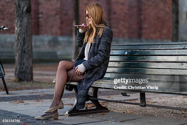 Sofia Grau sitting on a bench taking a photo with her phone for Instagram wearing a black plaid checked Urban Outfitters wool coat, a white creme...