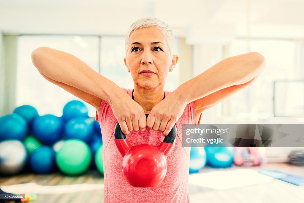 Mature Woman Exercising In Gym With Kettle Bell.