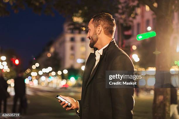 side view of man looking away while holding smart phone on city street at night - long coat stock pictures, royalty-free photos & images