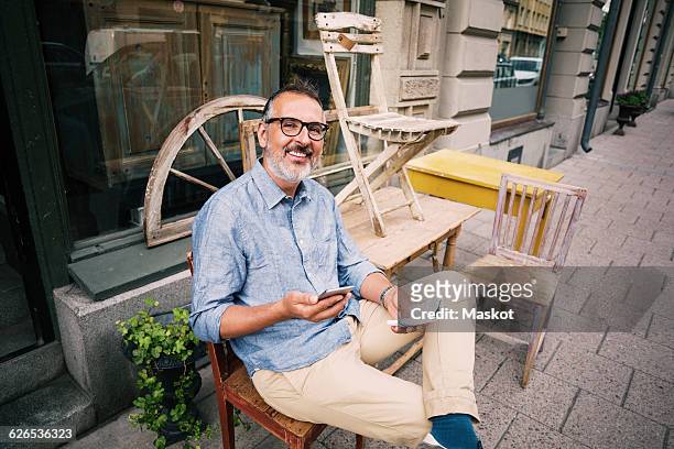 portrait of owner holding mobile phone while sitting outside antique shop - man in antique shop stock pictures, royalty-free photos & images