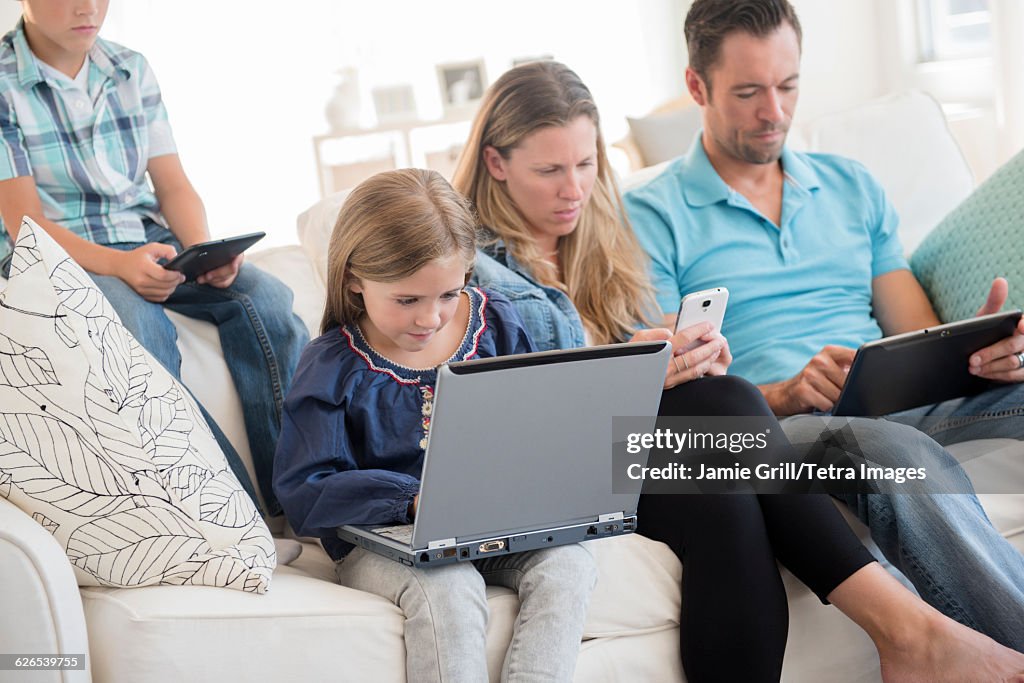 Family with two children (6-7, 8-9) sitting on sofa, using laptop and digital tablets
