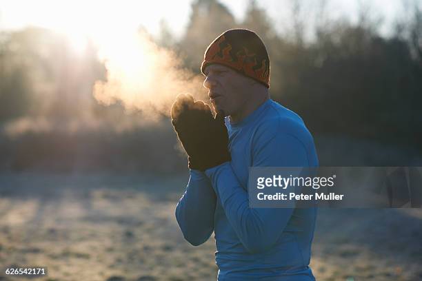 runner wearing knit hat and gloves, rubbing hands together, breathing cold air - cold morning stockfoto's en -beelden