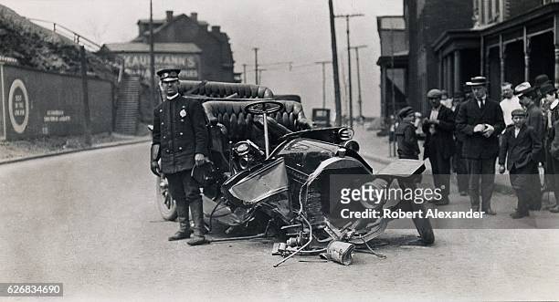 Police officer stands beside a circa 1905 automobile involved in an accident as a group of onlookers gathers nearby. Photograph is by Albert Mecham...