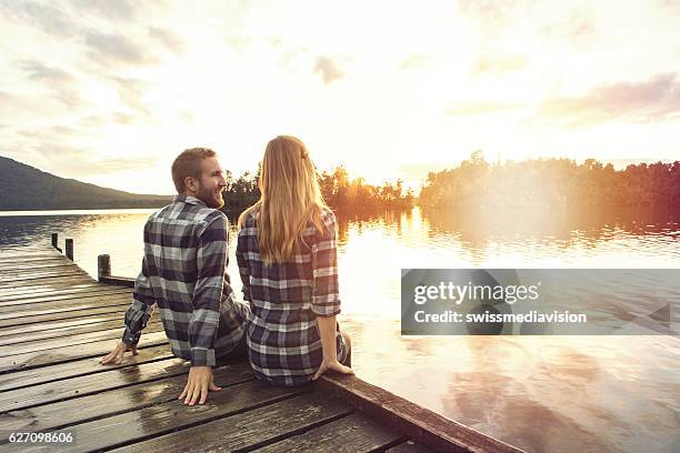 young couple sitting on lake pier enjoying sunset - bergsteiger stockfoto's en -beelden