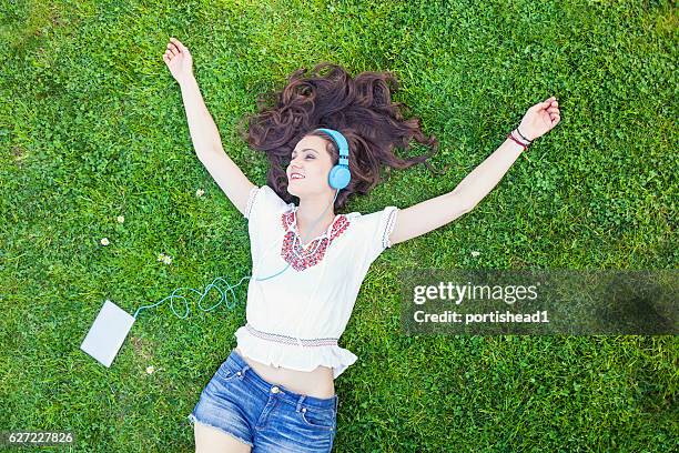 carefree young woman lying down on grass and listening music - girl lying down stock pictures, royalty-free photos & images