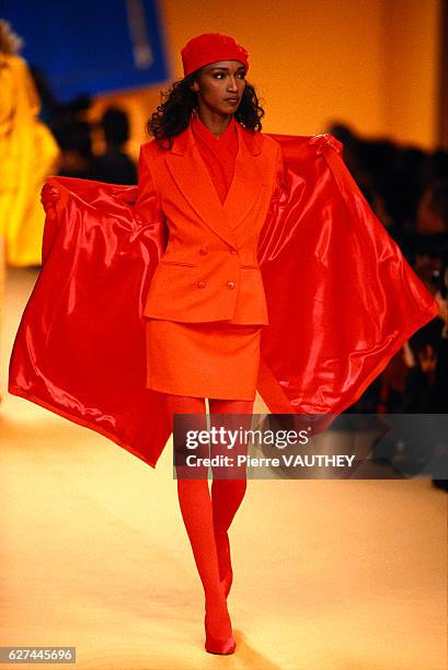 Fashion model wears a bright orange, ready-to-wear suit with matching coat, beret, and stockings by Japanese fashion designer Kenzo. She modeled the...