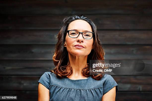 reife geschäftsfrau steht im büro gegen eine holzwand - female spectacles stock-fotos und bilder